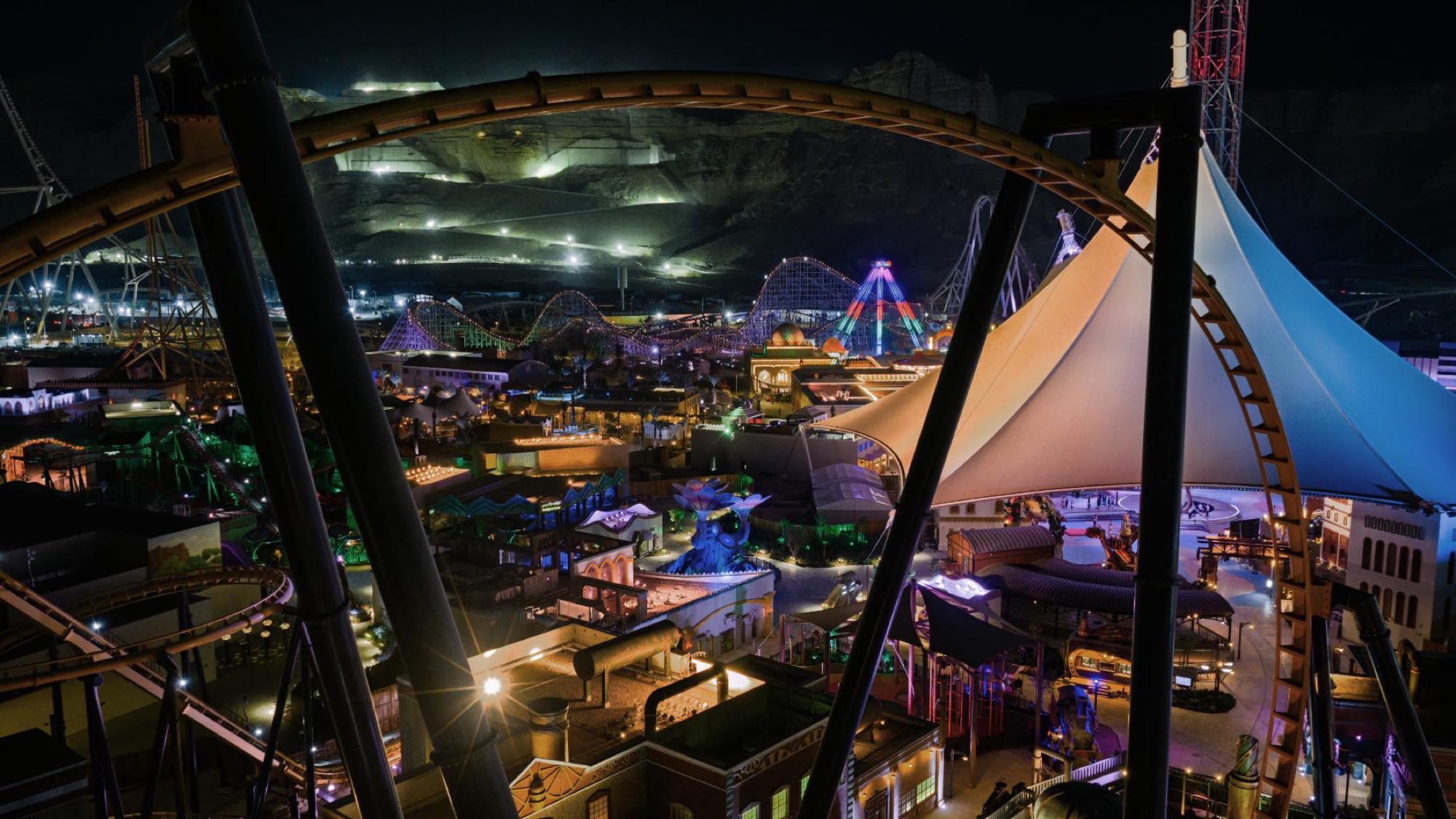 The open park at night, six lit lands seen through the loops of a coaster