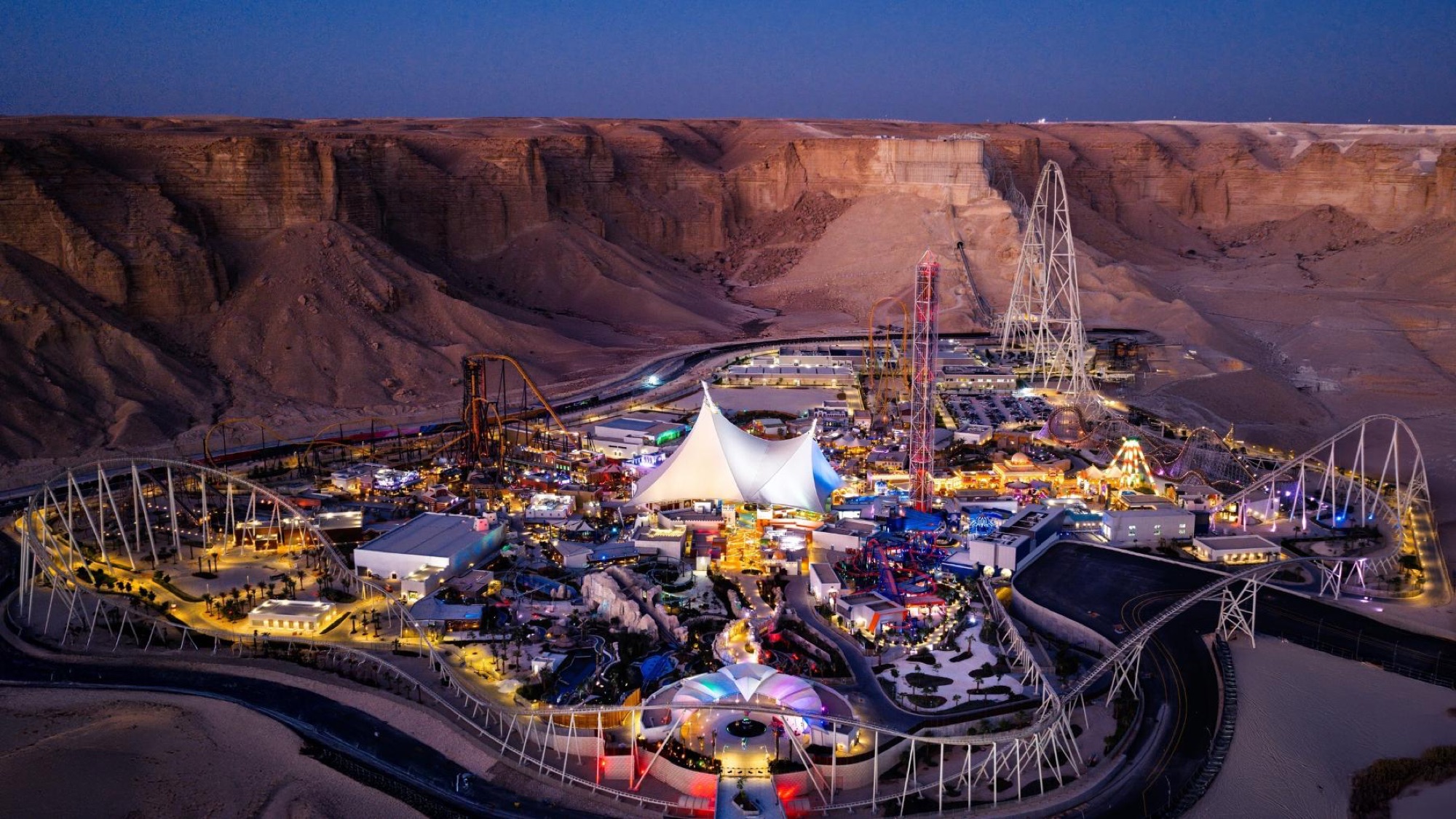 Six Flags Qiddiya City lit up at dusk beneath the Tuwaiq cliffs